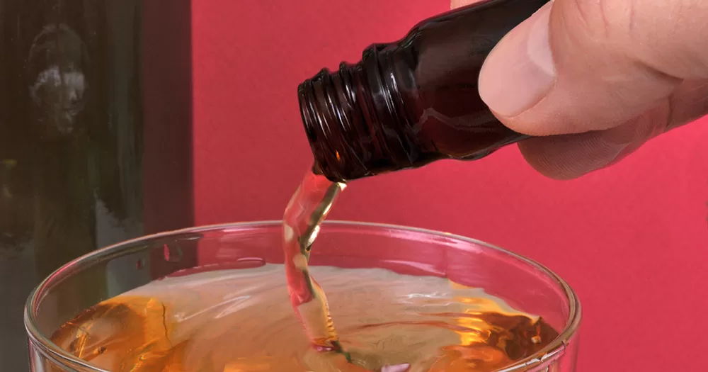 An image of liquid G, a drug used during chemsex that can cause addiction issues. the image shows a hand pouring orange liquid from a small brown glass bottle into a petri dish.