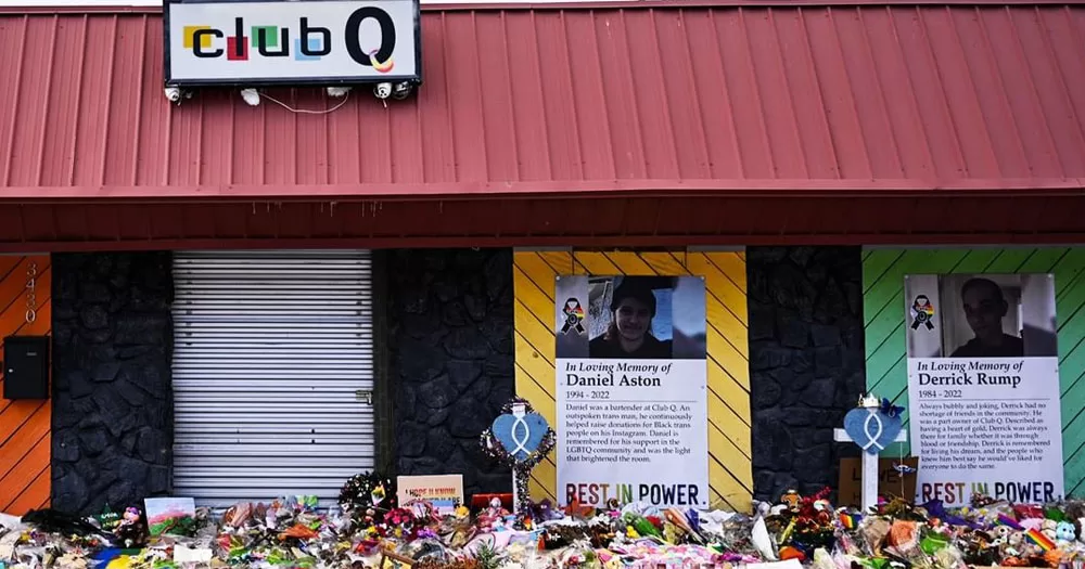 The image shows the front of Club Q where 5 people were killed in a hate crime shooting. The building is single story with a red metal roof. a neaon sign reading "Club Q" is mounted to the roof. In front of the building are bunches of flowers. On the wall are posters of the victims of the shooting.