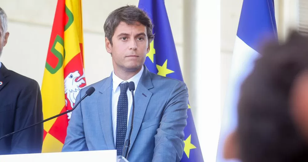 Image of Gabriel Attal standing at a podium. He wears a blue suit and stands in front of an EU and French flag.