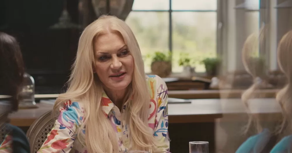 The image shows Rebecca Tallon De Havilland in the series Second Chances. In it a woman sits at a table. Her head is slightly tilted as she listens to a person off camera. She is sitting in a room in front of a window. She has long blonde hair which she is wearing down and a white blouse with a colourful floral pattern.