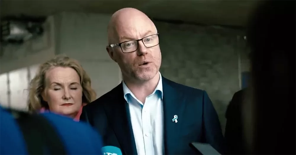 Stephen Donnelly speaking to reporters. He is photographed from the chest up, and wears a navy suit and glasses. I woman stands behind him at his right shoulder.