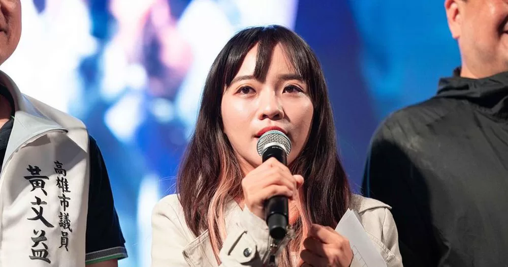 The image shows Huang Jie, the first openly LGBTQ+ legislator elected to parliament in Taiwan. In the photograph she is holding a microphone to her mouth. She is wearing a light grey jacket and has long black hair and red lipstick.