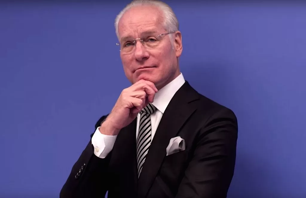 Photograph of Tim Gunn standing in a suit in front of a purple background