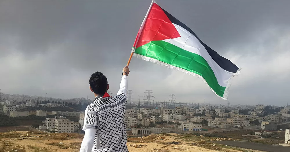 This article is about Irish LGBTQ+ organisations issuing an open letter calling for a ceasefire in Gaza. The image shows a man waving a Palestinian flag over his head in front of the Jordan skyline.