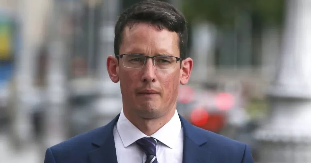 The image shows Enoch Burke who will remain in prison following yesterday's court hearing. It is a head and shoulders closeup taken in an outdoor setting. He is wearing a navy blue suit and tie, and a white shirt. He has a sullen look on his face and is wearing glasses.