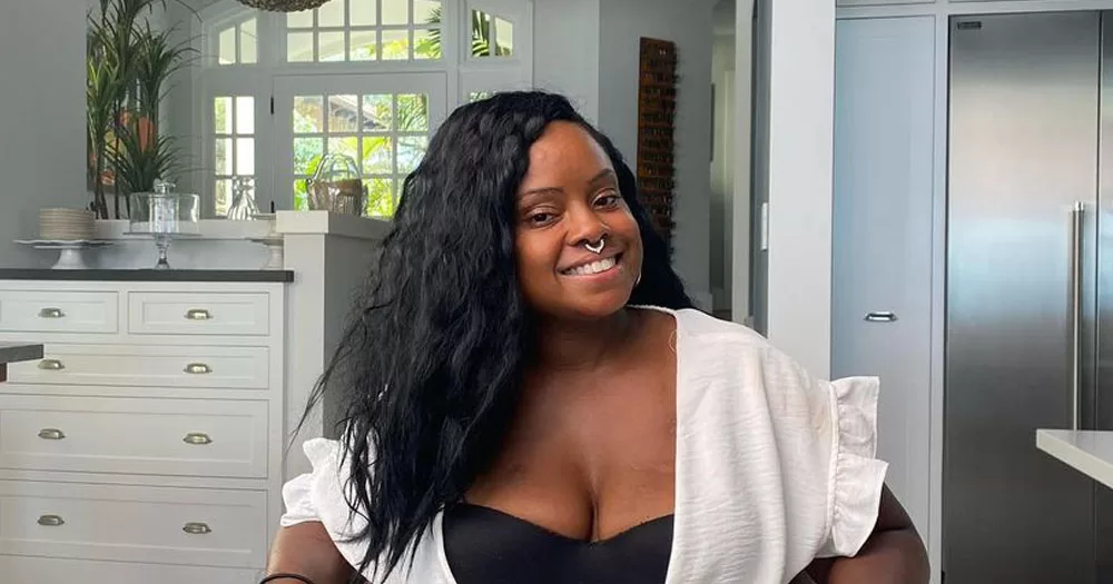 The image shows HIV and AIDS activist Hydeia Brodbent, who passed away suddenly. In the image she is sitting with her head tilted to the right. She is looking directly at the camera smiling. She has long wavy black hair. She is wearing it down, over her right shoulder. She is wearing a low-cut black bodice with a short-sleeved white light jacket over it.