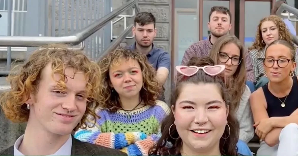 The image shows a group of young people sitting on steps outside a building. In the front left of the image is Joe Drennan who's classmates paid tribute to him at an award ceremony. He is smiling and has short curly blonde hair. He has a ring in his nose. To his left is a girl with hoop earrings and heart-shaped sunglasses resting on her head.