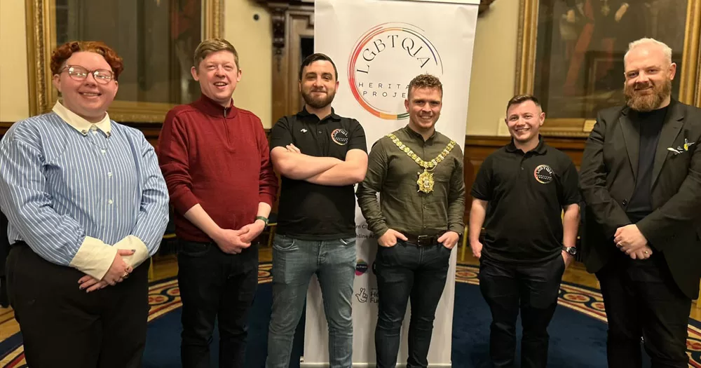 The picture shows a photograph of six people taken at the launch of a new LGBTQIA+ Heritage Project. From left to right are, Cara McCann, Director of HERe NI, Mukesh Sharma, The National Lottery Heritage Fund Northern Ireland NI Committee Chair (centre) with Scott Cuthbertson, Director of The Rainbow Project (left) and Adam Murray, Community Development Manager from Cara-Friend (right). They are standing in front of a pull-up banner with the LGBTQIA+ Heritage Project logo.