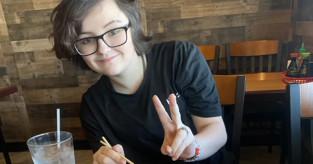 An image of Nex Benedict. They smile at the camera and hold up a peace sign with their fingers. They wear a black t-shirt and black-rimmed glasses.