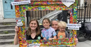 The image shows activist Ranae Von Meding with her wife and two of her children. In this article Ranae outlines why she will vote yes in the upcoming referendums. In the image, the family are posing behind a life-sized multicoloured picture frame. The frame has signs on it which read "love is love", "Still Not Equal" and "Family is Everything.