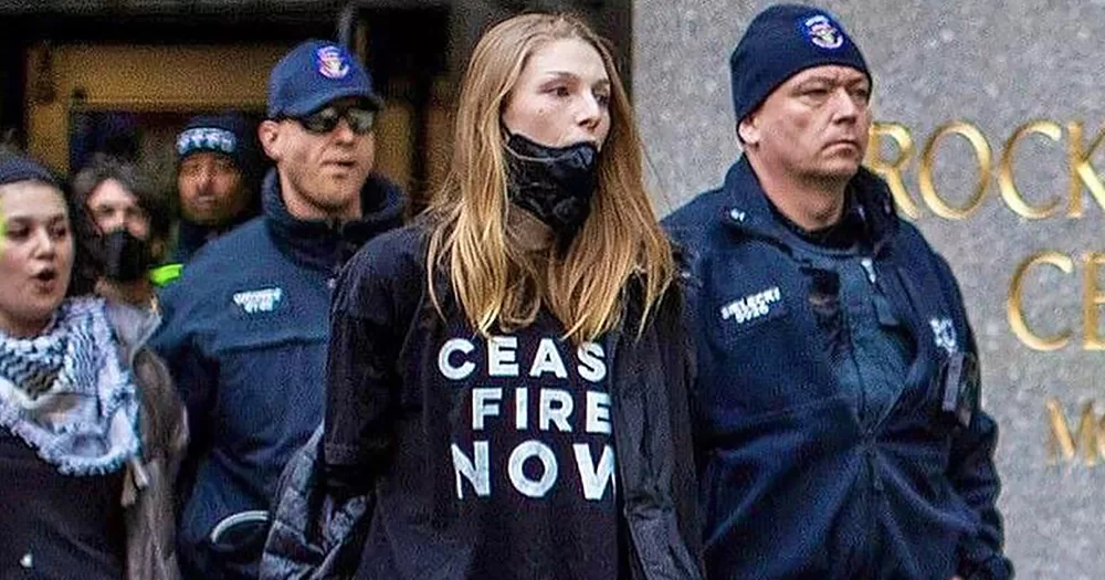 Actor Hunter Schafer being arrested for a pro-Palestine protest in New York. she is escorted out by police officers while wearing a mask and a t-shirt that reads "ceasefire now".