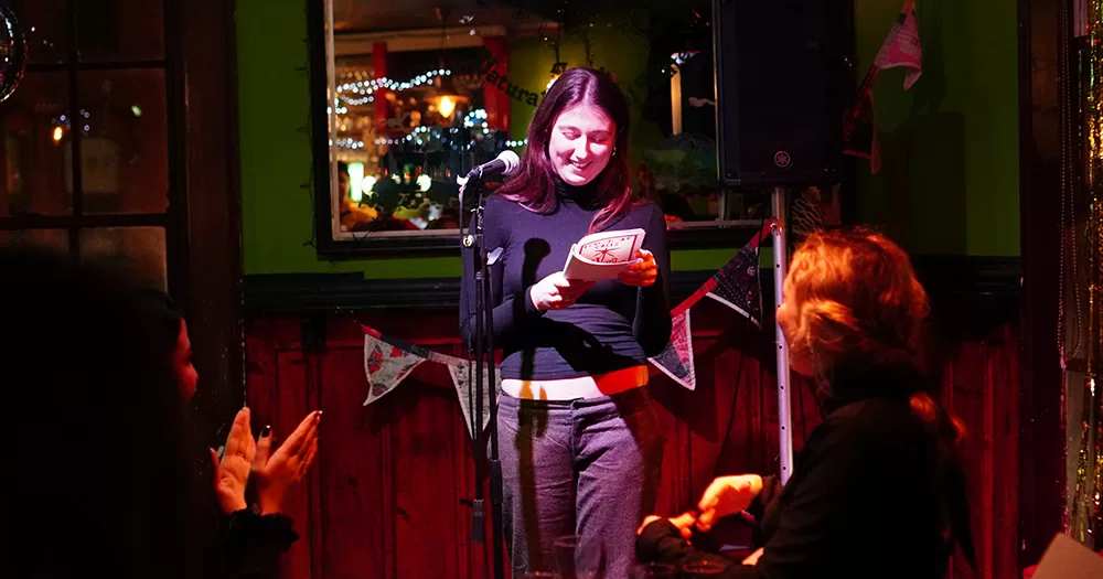 A person reading at a Lesbian Art Circle event. They stand at a microphone reading from the first issue of the publication.