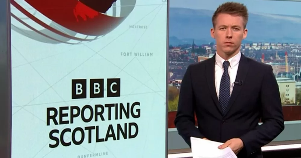 Journalist Nick Sheridan standing seriously beside a "BBC Reporting Scotland" sign. He is photographed from the waist up wearing a suit.