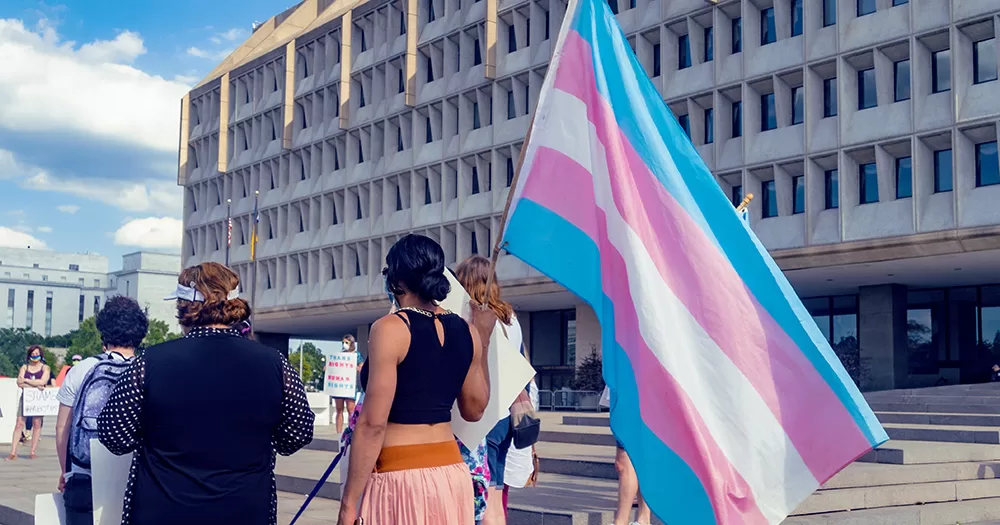 This article is about an increase in transphobic rhetoric in the EU. The image shows a small group of people walking in front of a large building, one of whom carries a large trans flag.