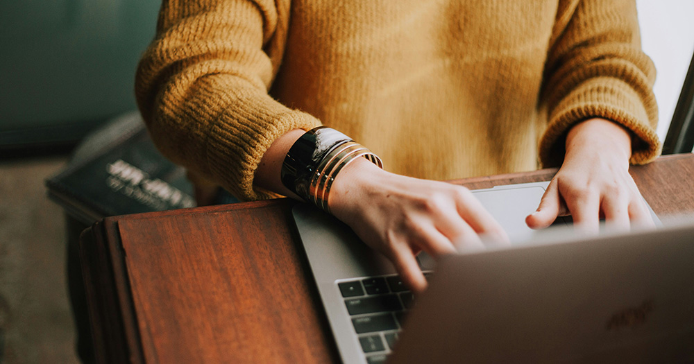 This article is about an LGBTQ+ homelessness survey. The image shows a person sitting at a wooden desk, using their laptop.