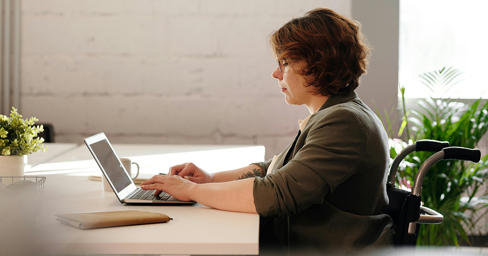 This article is about an online platform that connects LGBTQ+ people with disabilities. The image shows a person in a wheelchair sitting at a desk on their laptop.