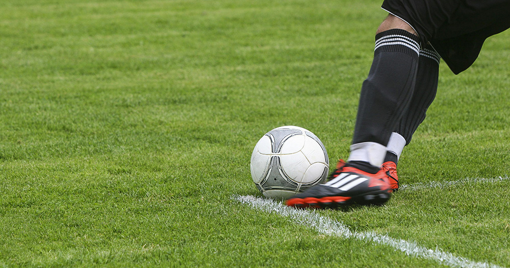 This article is about the National LGBT+ Helpline being promoted at League of Ireland matches. The image shows a person kicking a football on a grass pitch. The person is photographed from the knee down.