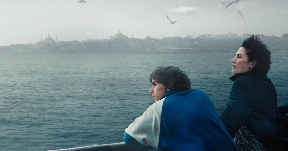 A photo from trans film Crossing. It shows two people leaning over a railing, looking out at sea as gulls fly above them.