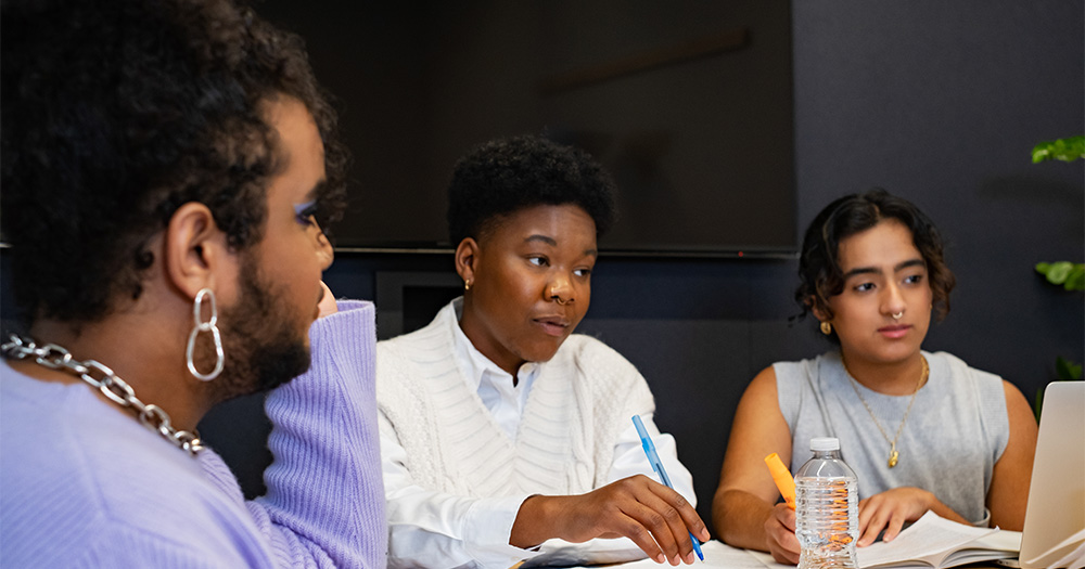 People, including LGBTQ+ professionals, taking part in a work meeting. They are sitting around a table and taking notes.