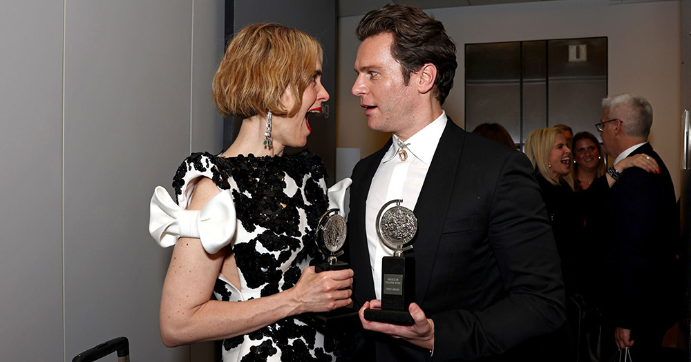 LGBTQ+ Tony Award winners Sarah Paulson and Jonathan Groff pose for a photo with their trophies. They are photographed from the waist up, facing each other with their mouths open.