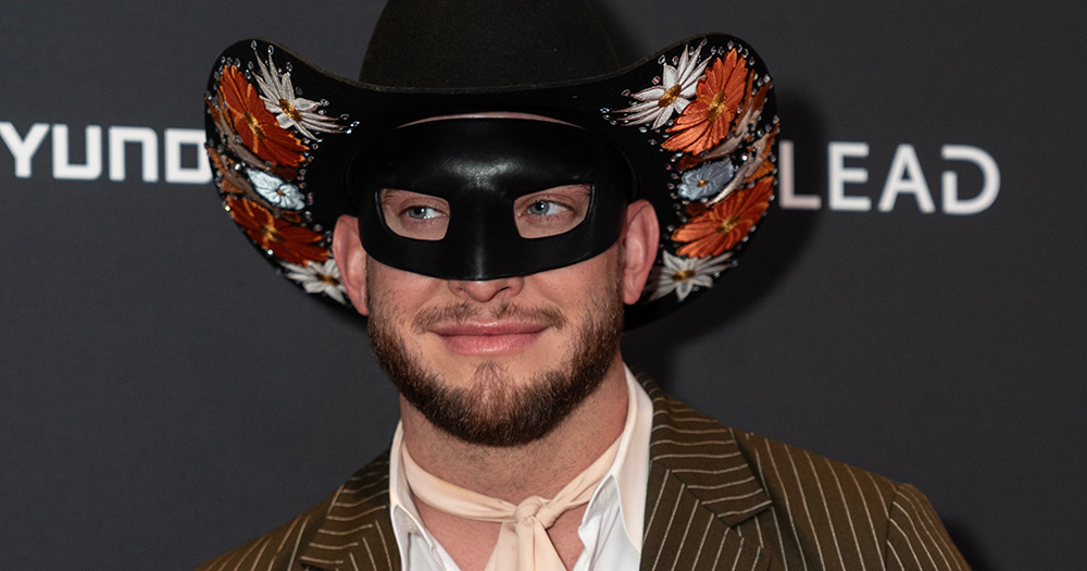 Close up image of Paper Magazine cover star Orville Peck. He wears a cowboy hat and eye mask and smiles with his mouth closed.