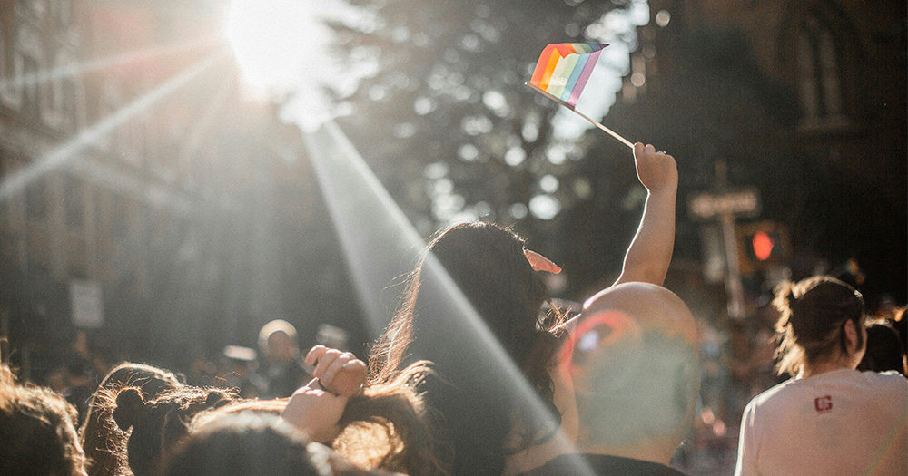 A girl stands on someone's shoulders during a Pride parade to hold up a mini Pride flag.