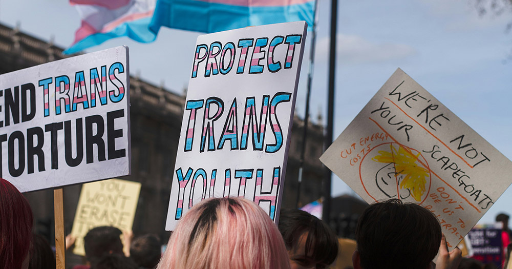 This article is about gender-affirming healthcare in Florida. The image shows a group of people holding pro-trans signs in front of a building.