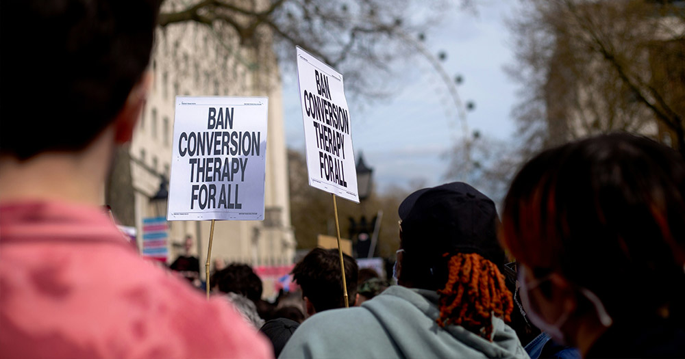 A group of people standing around holding signs that read: "Ban Conversion Therapy For All".