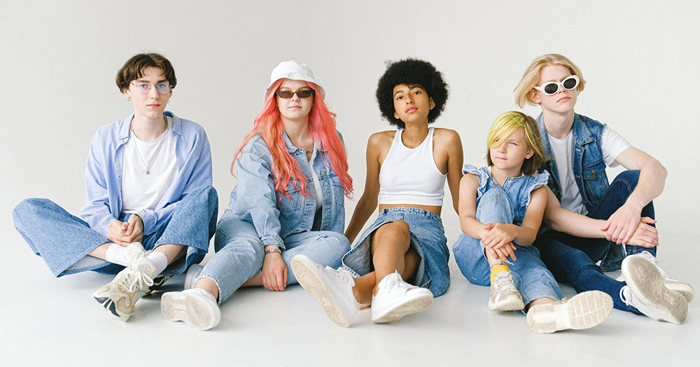 A group of five adolescents with various skin tones and hair colours. They are wearing variations of gender-neutral fashion like white tops and light-blue denim ensembles.