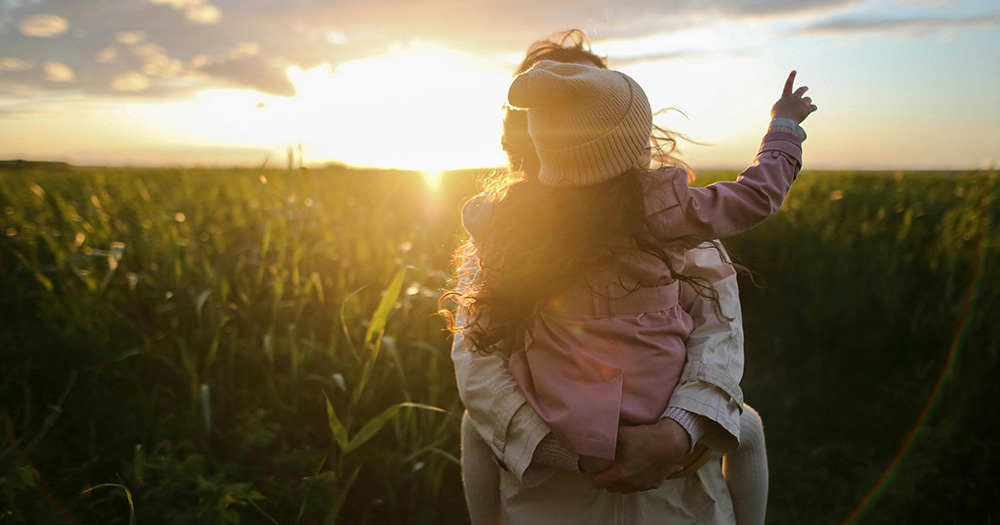 This article is about same-sex parents being unequal under Irish law. The image shows a mother holding a child in a green field. The sun is setting in the background.