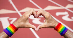 This article is about a documentary on LGBTQ+ inclusion in sports. In the photo, hands holding up a heart in front of a running track. Pride flag sweat bands are worn around the wrists.