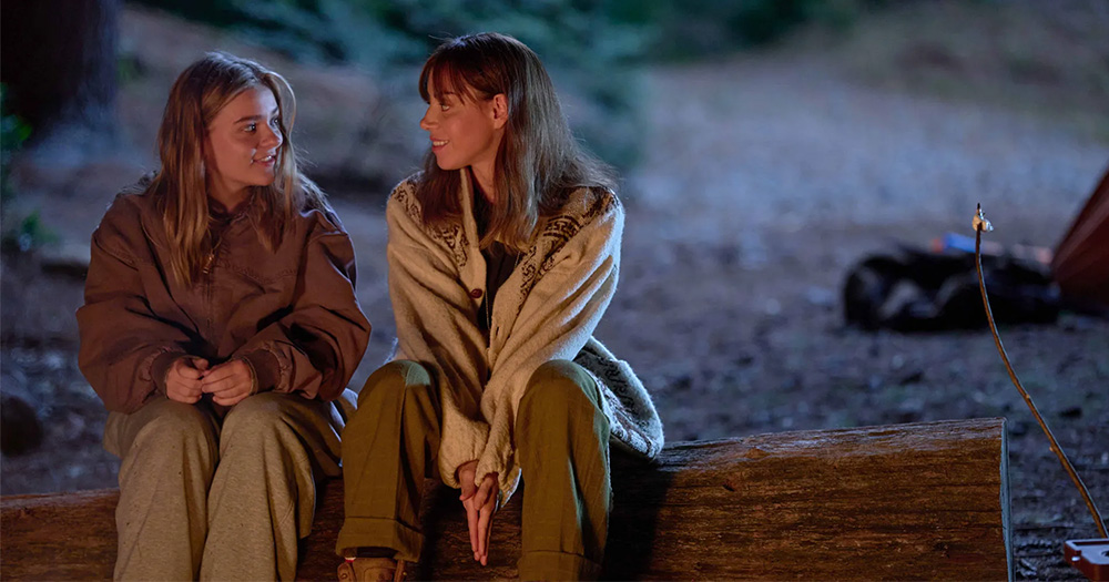 Teen Elliott (​​Maisy Stella) and adult Elliott (Aubrey Plaza) sit next to each other on a log while talking. Taken from the film My Old Ass.