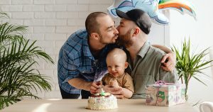 This article is about the new Assisted Human Reproduction law and how it impacts LGBTQ+ families. The image show two gay dads kissing on the cheek while their baby sits and smiles in front of them, with a birthday cake on the table.