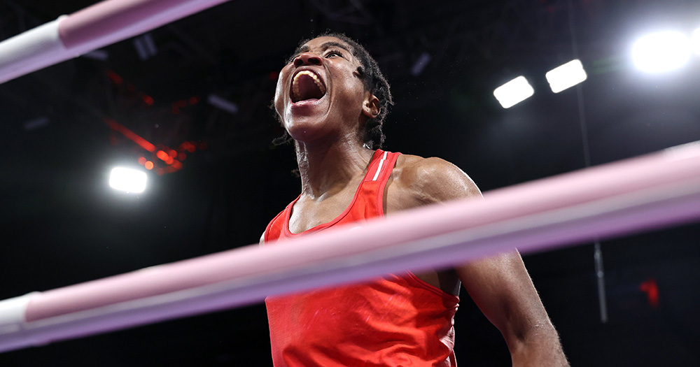 Image of boxer Cindy Ngamba. She is photographed through the ropes of the ring, cheering and wearing red boxing attire.