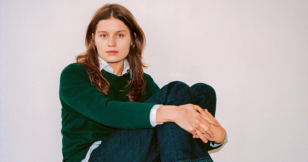 girl in red sitting down holding her knees to her chest. She wears a green jumper with a shirt underneath and blue jeans.