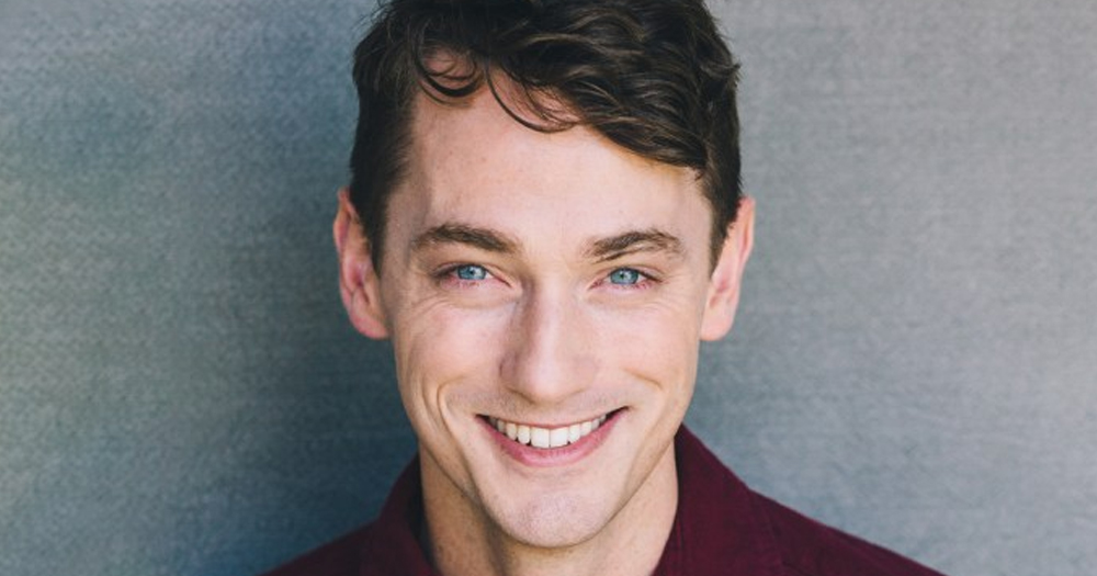 Photo of Michael Thomas Grant, who has just been cast as a new gay character in Grey's Anatomy, smiling at the camera on a grey background.