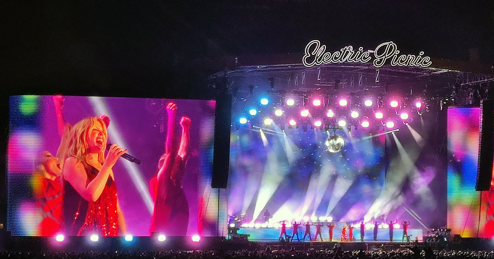 Kylie Minogue performing at Electric Picnic, with a crowd watching her on the stage with purple and blue lights and a big screen on the left.