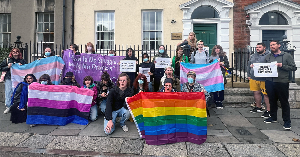 Activists gathered in front of Sinn Fein's headquarters in Dublin for a protest. They are posing with trans flags, Pride flags and other flags, as well as signs with messages of support for the trans community.