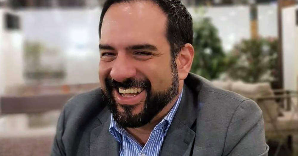 Manuel Guerrero Aviña, a gay man who was arrested and convicted in Qatar, smiling at the camera, wearing a grey blazer and light blue t-shirt.