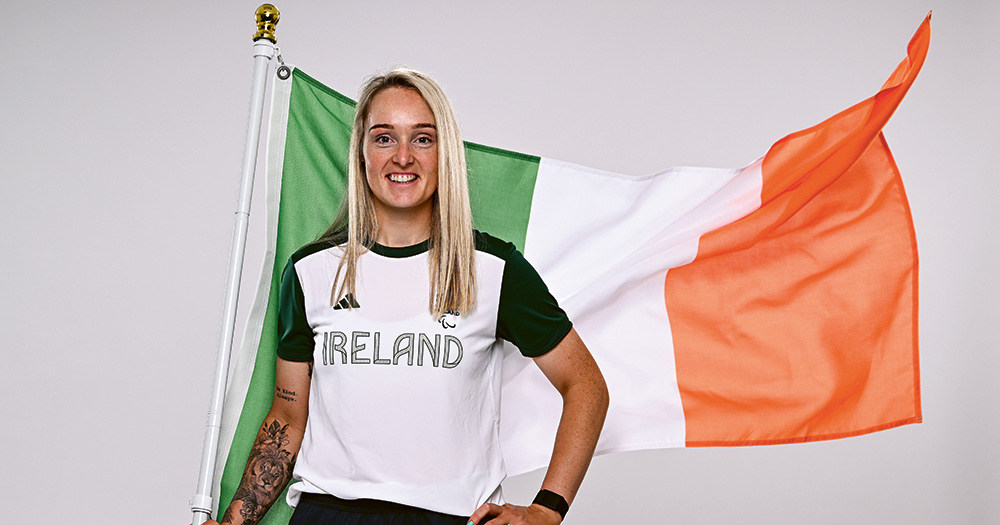 Richael Timothy posing in Team Ireland gear with a large Irish flag flying behind her.