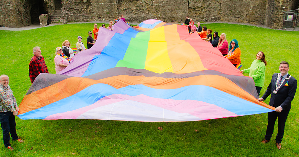 Promotional image for Roscommon Pride. Organisers and the Lord Mayor hold a large progress Pride flag outside on a grass area.