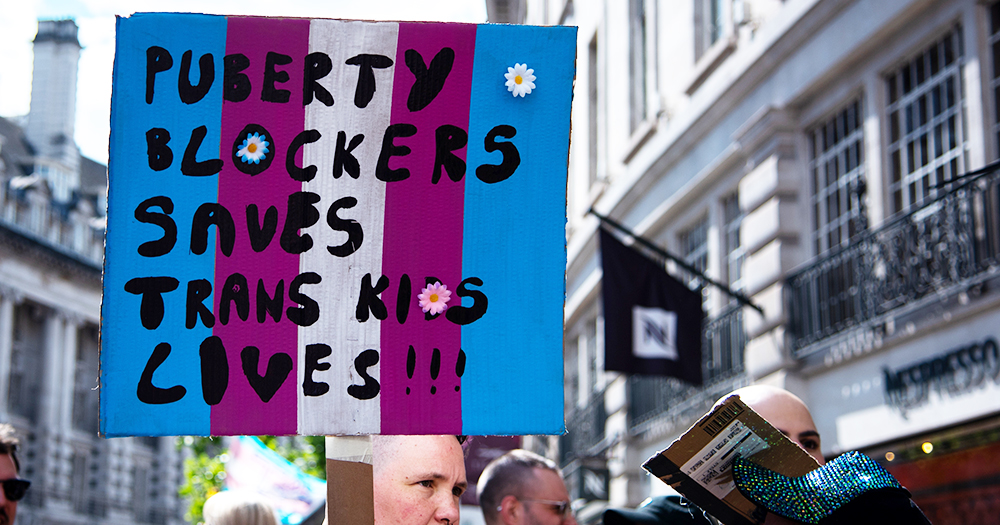 This article is about Sinn Fein backing a puberty blockers ban in Northern Ireland. In the photo, a person holding a banner that reads "Puberty blockers saves trans kids lives".