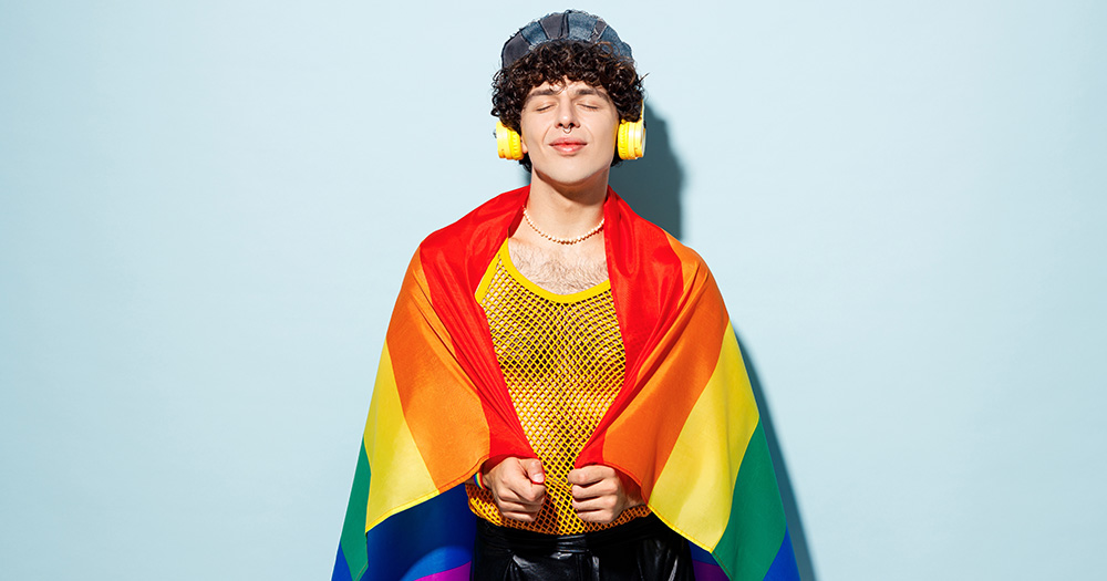 This article is about Irish queer podcasts. The image shows a person with a rainbow flag wrapped around them. They wear headphones and a hat.