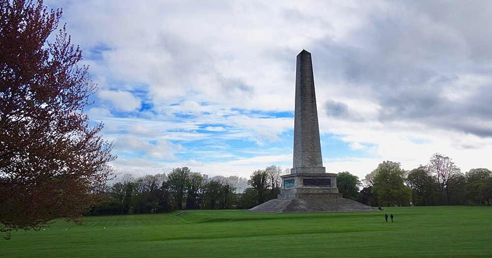 Photo of Phoenix Park, with a monument standing on a green field, with trees in the background and on the left.