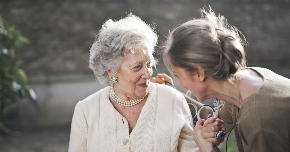 This is an article about LGBTQ+ retirement communities. Pictured are two women are looking at each other and are smiling and laughing. The woman on the left is old and wears a white woolen jacket. The woman on the right is a bit younger than her and holding her hand.