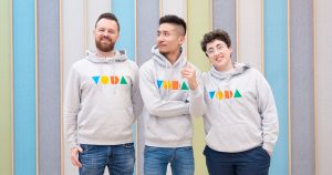 The team behind Trans Library, a mental health resource for trans people, with three people standing in front of a colorful wall wearing the same grey sweatshirt.