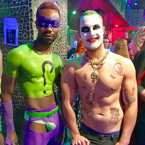 This queer couple costume for Halloween is about Riddler and Joker, so the two men pictured are wearing makeup to look like those two. One is wearing neon green and purple makeup, the other one white and green makeup.