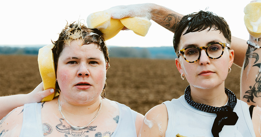 This is an interview with the band ARXX about their new album Good Boy and their upcoming show in Dublin. Pictured are bandmembers Hanni (left) and Clara (right) as they shot the video for their song Crying In The Carwash. Both are literally washed by other people who are shampooing them both with sponges just as cars in a carwash. Both are wearing white tops and are looking right into the camera.