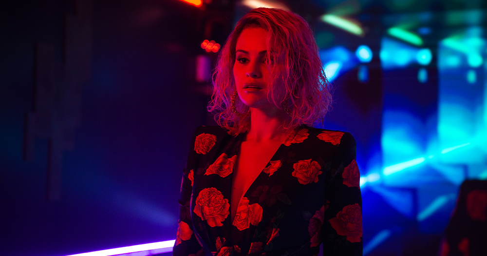 Still from film Emilia Perez, with Selina Gomez standing in a dark room with blue and red lights