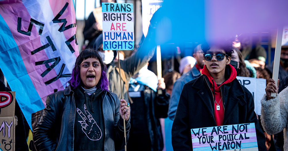 This article is about trans people being denied hormone replacement therapy in the UK. In the photo, people protesting for trans rights with trans flag and signs.